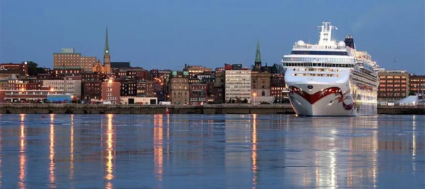 A large white cruise ship with red accents docked at a harbor next to a city skyline at dusk, with reflections visible on the water.