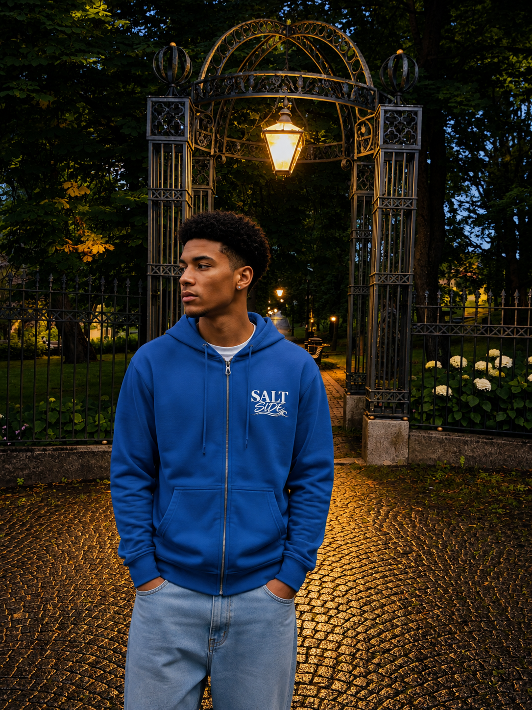 A man wearing a royal blue zip-up hoodie with 'SALT' printed in white on the chest, standing in a park in front of an illuminated metal garden arch at dusk.